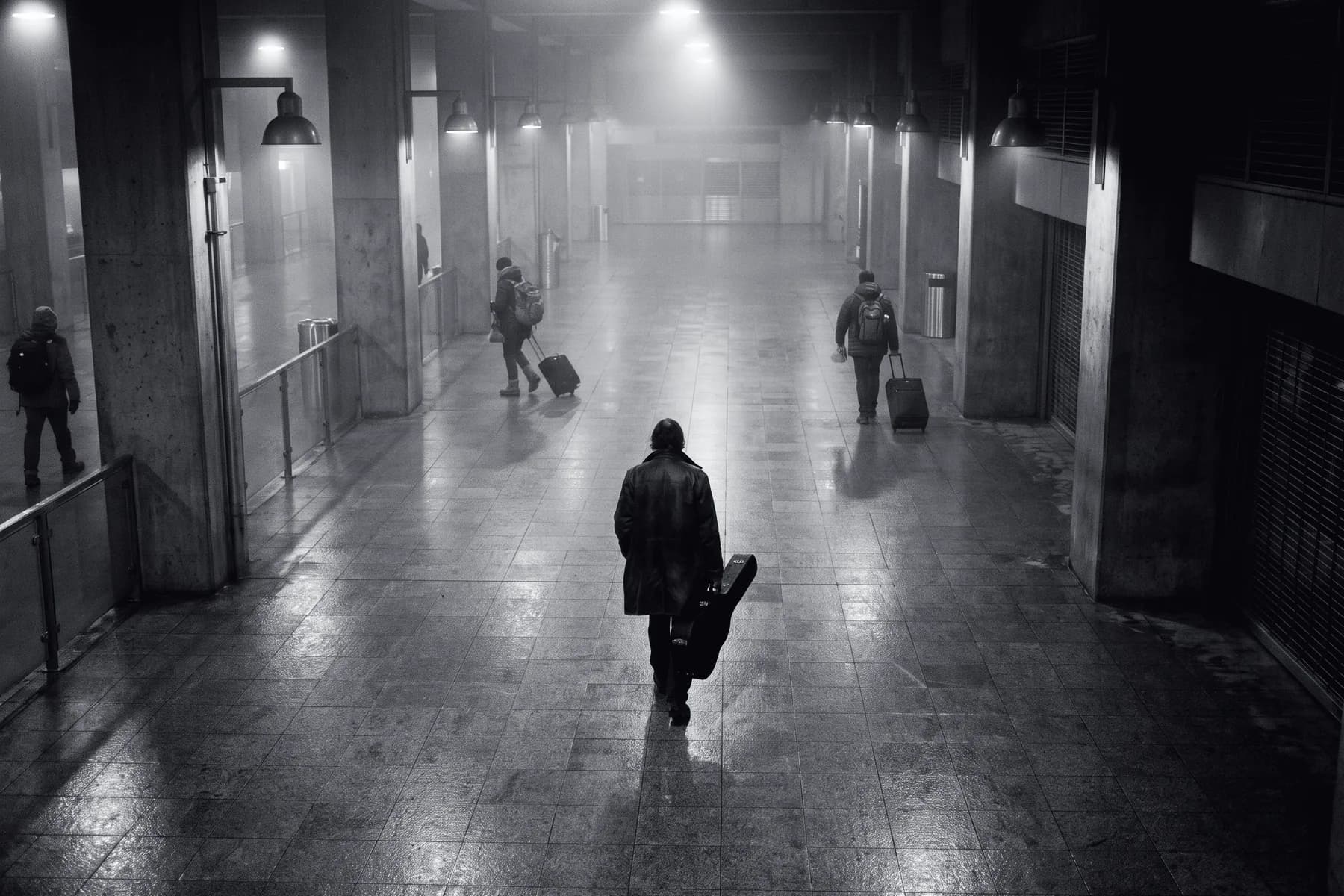 Illustrative frame: a transport concourse at night from an elevated camera position. One person walking alone carries a long rigid object held low against their leg. Shown in black and white, with the annotated regions revealed in colour.